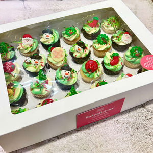Box of Christmas-themed cupcakes with green icing and red decorations, featuring the brand 'Bakealicious'.