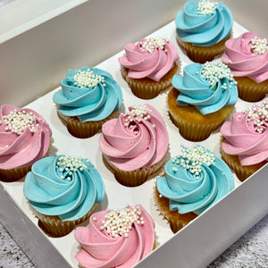 Twelve frosted cupcakes arranged in a white box, alternating between light pink and light blue swirled frosting, topped with white sprinkles.