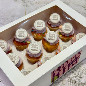 A white rectangular gift box with a clear plastic top window displaying multiple frosted cupcakes. Each cupcake is topped with a round white sticker printed with the Nesñus logo.