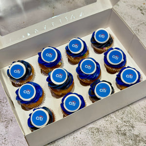 A white cardboard box containing ten cupcakes, each topped with blue frosting and a circular blue-and-white Citi Bank logo topper, photographed on a light stone-like surface.