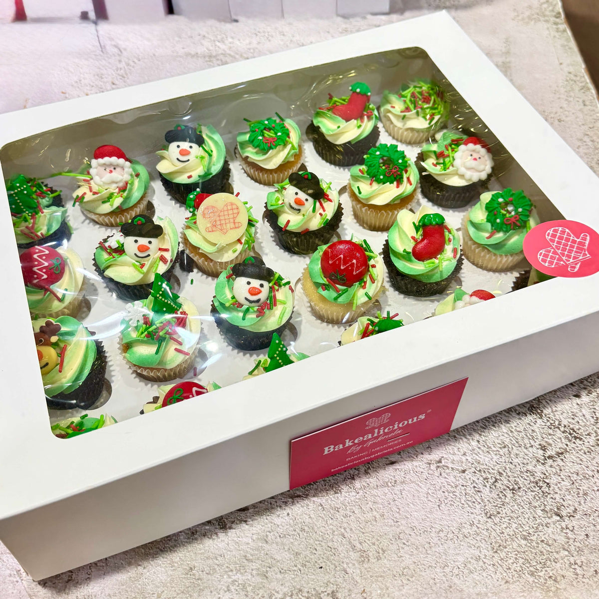 Box of Christmas-themed cupcakes with green icing and red decorations, featuring the brand 'Bakealicious'.