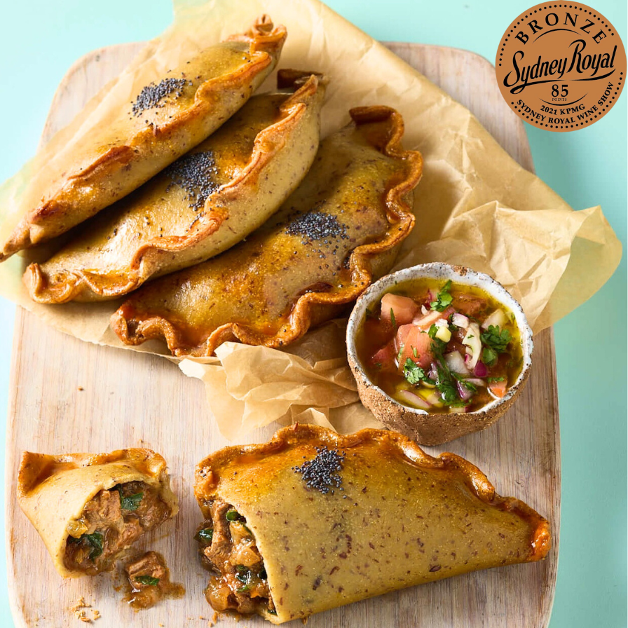 Three baked empanadas on a wooden board with a small bowl of salsa, photographed on a light blue background. A bronze Sydney Royal 55 award badge is visible in the top right corner.