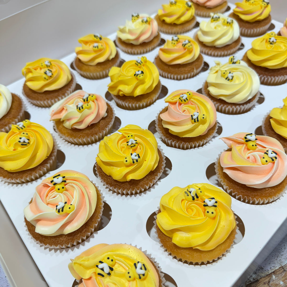 A tray of frosted cupcakes arranged diagonally. The cupcakes feature swirls of yellow, peach, and cream frosting, with some decorated to look like bumblebees.