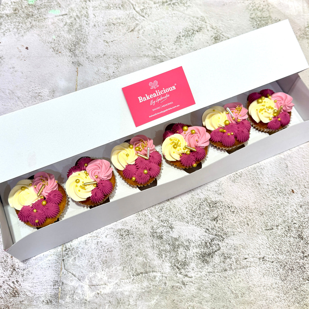 A white bakery gift box containing six decorated cupcakes with pink, cream, and yellow frosting, displayed on a textured gray surface. The inside lid features a pink label for 'Bakealicious By Gabriela'.