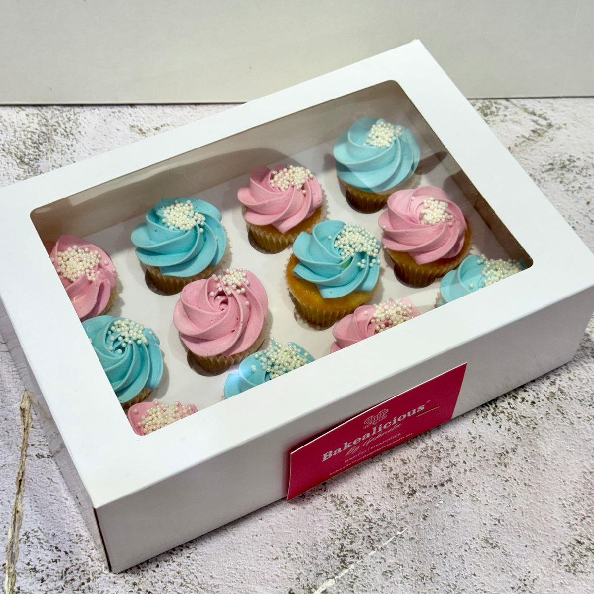 A white bakery gift box with a clear window displaying 12 frosted cupcakes, decorated with pink and light blue frosting and white sprinkles. A red label on the front of the box reads 'Bake Shop of Jones'.