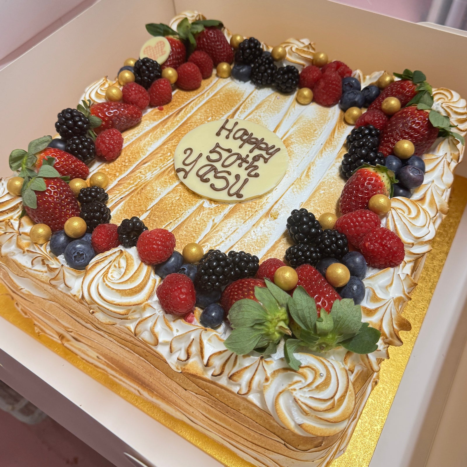 A square birthday cake with white meringue piped on the sides and top, decorated with fresh berries, gold-colored drizzle, and a central chocolate plaque reading 'Happy 50th YUSU!'.