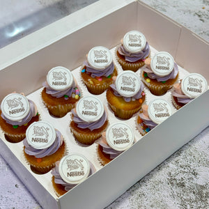 A white cardboard box containing multiple frosted cupcakes arranged in a tray. Each cupcake has light purple/lavender frosting, a golden-brown cupcake liner, and a round white Nestle topper on top.