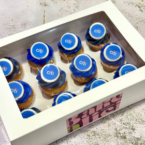 A white gift box containing several frosted cupcakes topped with blue icing swirls and circular blue-and-white Citi Bank logos, photographed on a light textured surface.