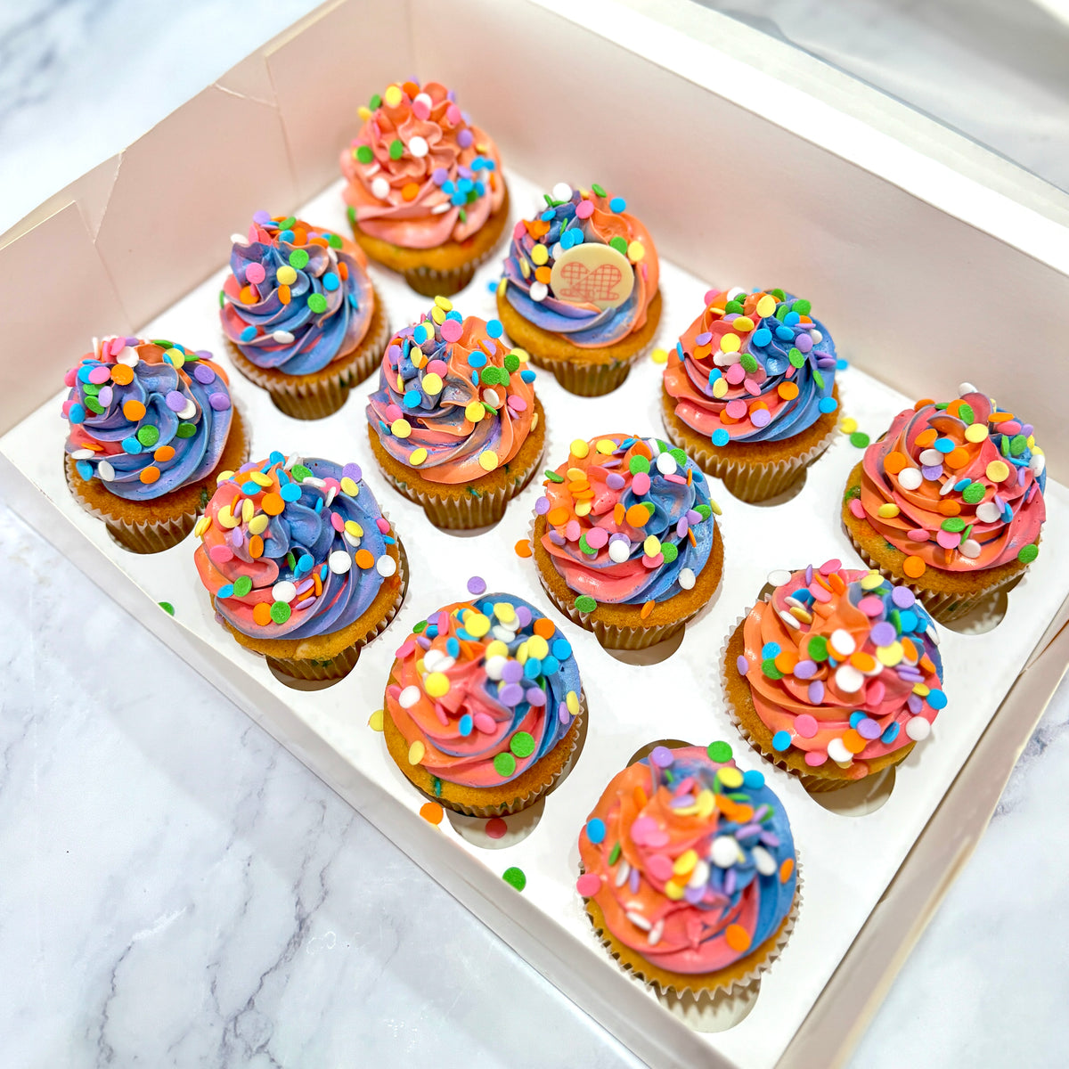 A set of 12 colorful frosted cupcakes arranged in a white bakery box on a light marble surface. The cupcakes have swirls of pink, orange, and blue frosting, topped with multicolored sprinkles, and one has a small heart-shaped topper.