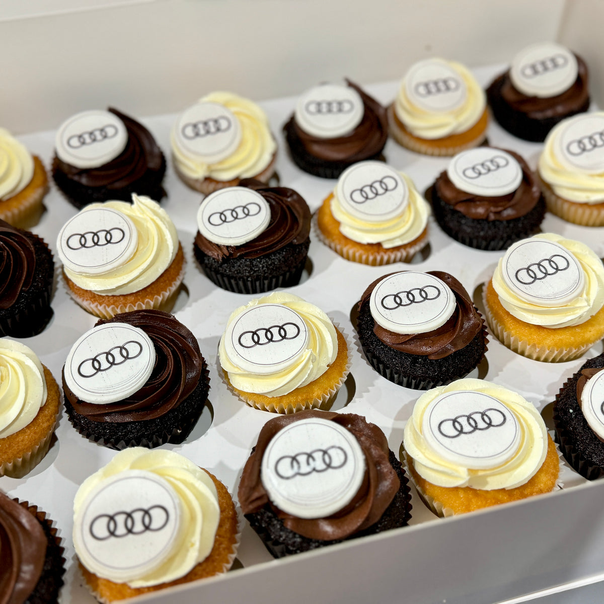 A box of cupcakes decorated with white and dark chocolate frosting, each topped with a white round piece printed with the four-ring Audi logo.