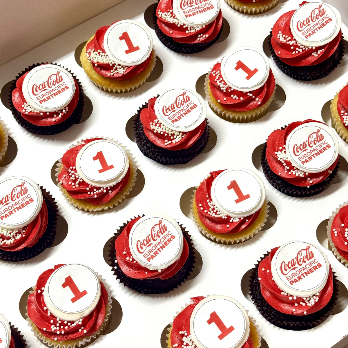 A box of Coca-Cola-themed 1st birthday cupcakes with red and white frosting, white toppers featuring the red Coca-Cola script and 'Coca-Cola Partners,' and sprinkles, arranged in a white cupcake tray.