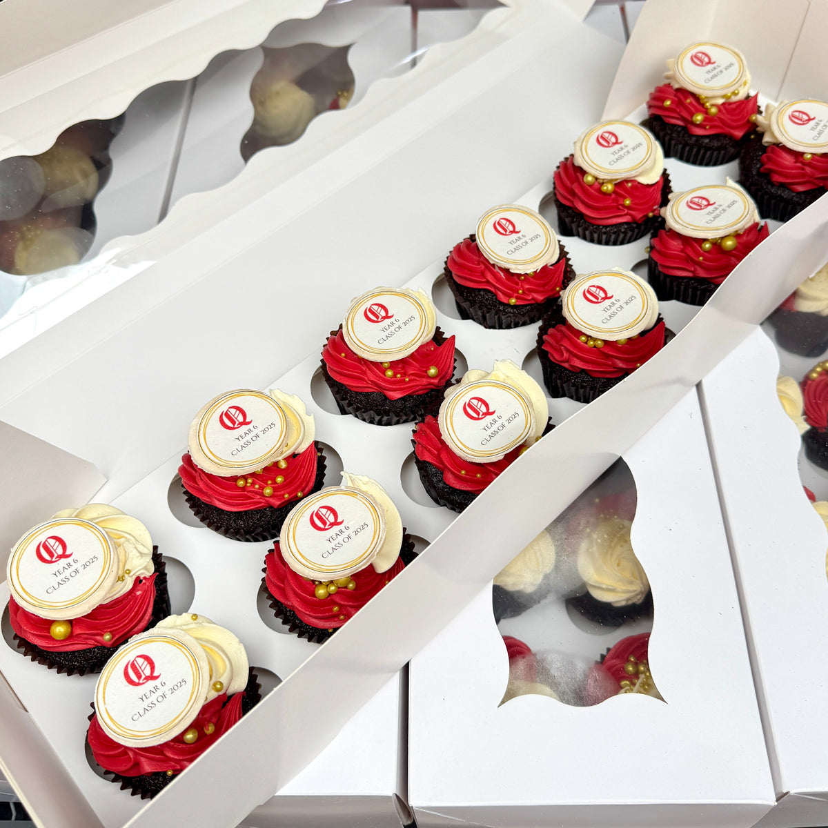 Open white bakery boxes containing red-frosted cupcakes topped with white round toppers printed with Queenwood School logo and the words 'Year6, Class of 2025'.