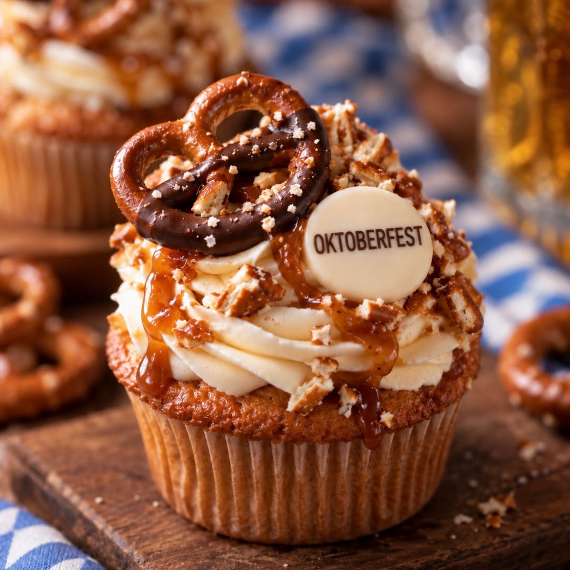 A close-up of a frosted cupcake topped with a pretzel, caramel drizzle, cookie crumbs, and a round white decoration printed with 'OKTOBERFEST'. The cupcake sits on a wooden board with more pretzels and a blue-and-white checkered cloth in the background.